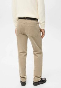 Pantalones chinos beige de corte slim, con dos bolsillos traseros y textura suave, combinados con zapatos negros. Fondo blanco liso.
