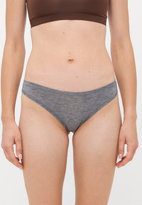 Vrouw in een bruine crop top en grijze low-rise onderbroek, staand tegen een eenvoudige witte achtergrond met de handen ontspannen naast zich.