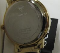 Montre en acier inoxydable plaqué or avec logo "GUESS" gravé, étiquette indiquant qu'elle est résistante à l'eau et numéro de modèle. Surface lisse et brillante.