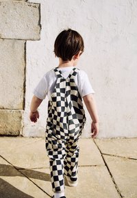 Salopette a scacchi bianca e nera in cotone, indossata sopra una t-shirt bianca, con spalline regolabili e tasche, vista da dietro.