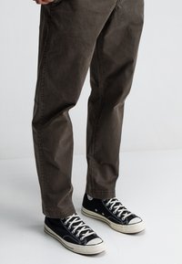 Donkerbruine broek met een relaxed fit, voorzien van zijzakken. Gecombineerd met zwarte canvas sneakers met witte zolen en veters.