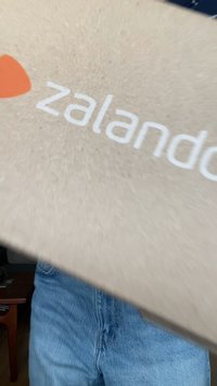 Barna kartondoboz, fehér és narancssárga Zalando logóval, részben eltakarva, farmernadrág mellett egy fa felületen.