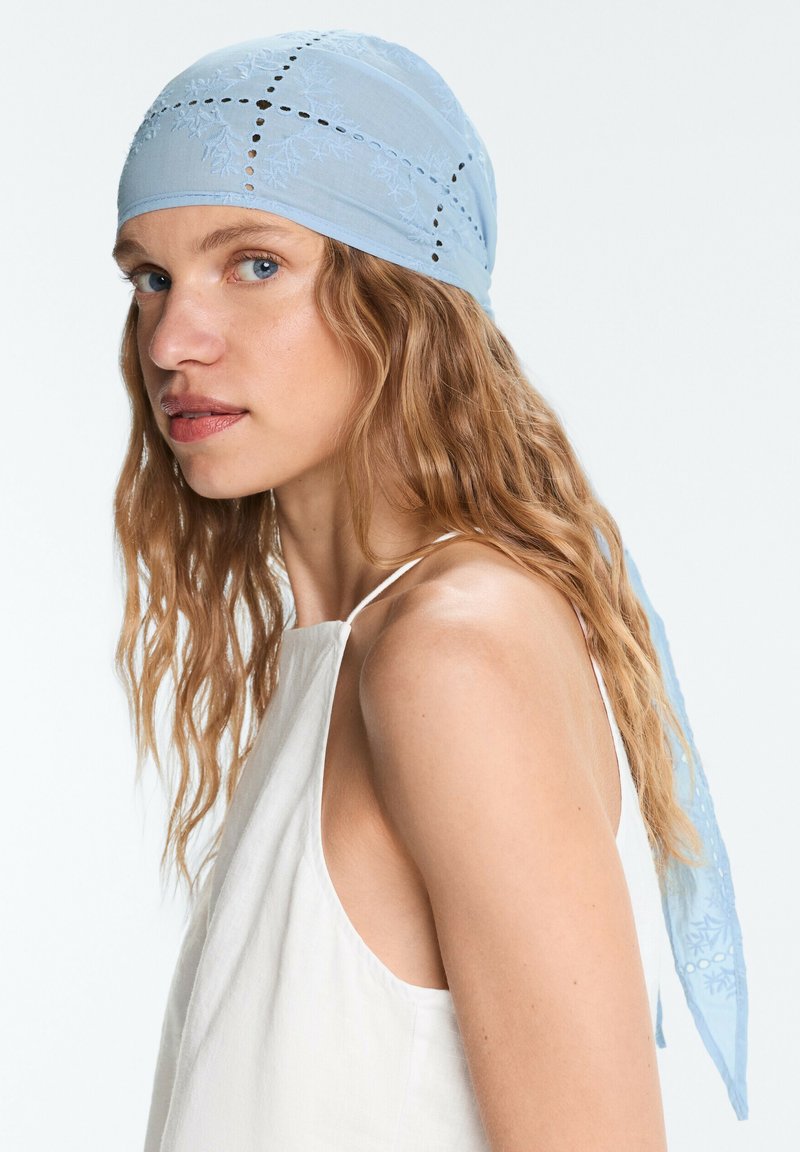 Femme aux cheveux blonds ondulés portant un foulard brodé bleu clair et un haut blanc sans manches, regardant légèrement sur le côté.