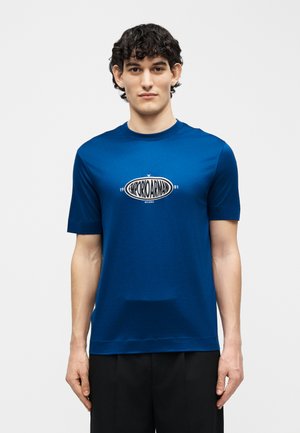 Άνδρας με κοντά σγουρά μαλλιά που φοράει μπλε μπλουζάκι Emporio Armani και μαύρο παντελόνι, στέκεται μπροστά σε απλό φωτεινό φόντο.