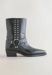 La bota de tobillo de cuero negro presenta remaches metálicos decorativos, una correa con una hebilla circular y un tacón bajo en bloque con un diseño de punta afilada.