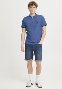 Blå polo skjorte med krage og tre knapper, kombinert med denimshorts og hvite sokker. Skoene har svarte striper og grå detaljer.