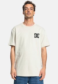 Hellbeige Baumwoll-T-Shirt mit lockerer Passform, ausgestattet mit einem schwarzen DC-Logo auf der linken Brustseite und kurzen Ärmeln.