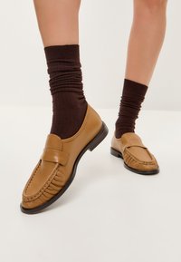 Bruine leren loafers met een ronde neus, voorzien van een voorkraag en geplooide details, gecombineerd met bruine geribde sokken.