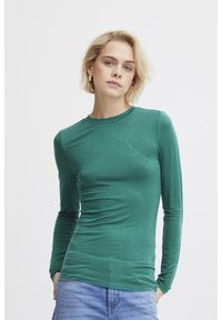 Långärmad grön skjorta i mjukt tyg, med rund halsringning och smal passform som har en mjuk textur. Bärs med blå jeans.