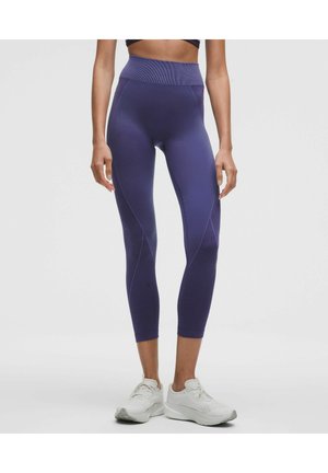 Žena nosící fialové texturované legíny s vysokým pasem, zkrácené nad kotníky, a bílé sportovní boty, stojící před jednobarevným pozadím.