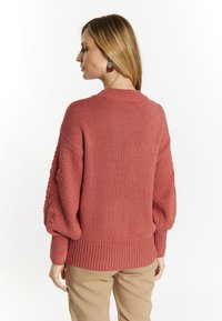 Różowy sweter z dzianiny z teksturowanymi balonowymi rękawami i ściągaczami, połączony z jasnobeżowymi spodniami. Widok z tyłu.