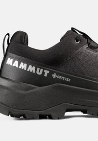 Μαύρο πεζοπορικό παπούτσι Mammut με υφασμένο πάνω μέρος, παχιά σόλα, λογότυπο Gore-Tex, θηλιά στο πίσω μέρος και σταθερά κορδόνια σε γκρι φόντο.