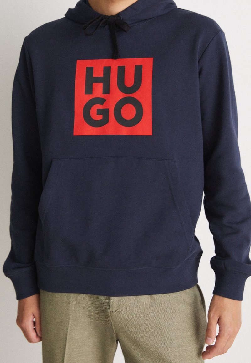 Persona con sudadera azul marino con un rectángulo rojo y texto negro "HUGO" en el pecho, combinada con pantalones beige claros.