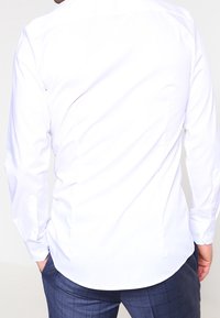 Man som bär en figursydd vit långärmad skjorta och mörkblå rutiga byxor, står med händerna i fickorna, bakifrån.