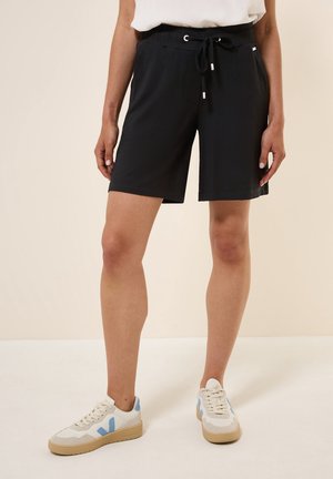 Femme portant un short noir à cordon de serrage et des baskets blanches avec des accents bleus, debout sur un sol et un mur beige clair.