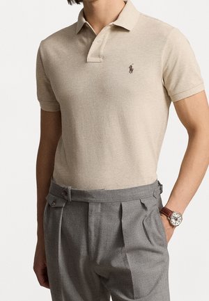 Beżowa koszulka polo z krótkim rękawem, kołnierzykiem i listwą na guziki, zestawiona z wysokim stanem szarych plisowanych spodni i zegarkiem na rękę.