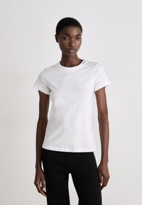 T-shirt bianca in cotone con maniche corte, scollo rotondo e dettaglio del logo sulla spalla. Abbinata a pantaloni neri a coste.