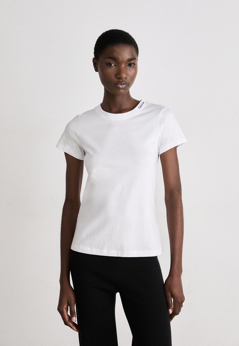 T-shirt bianca in cotone con maniche corte, scollo rotondo e dettaglio del logo sulla spalla. Abbinata a pantaloni neri a coste.
