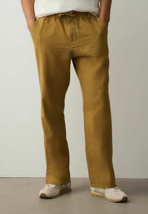 Mann trägt senffarbene gerade Hose mit Händen in den Taschen und weißen Sneakers, steht vor einem schlichten hellen Hintergrund.