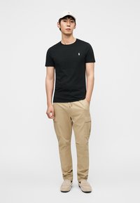 Μαύρο t-shirt με κοντό μανίκι, χακί cargo παντελόνι και μπεζ μοκασίνια. Το t-shirt έχει μικρό λογότυπο· τα cargo παντελόνια διαθέτουν πλαϊνές τσέπες.