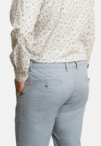 Pantalones azul claro con un acabado texturizado, que presentan bolsillos traseros con cierres de botón. El modelo lleva una camisa de patrón floral.