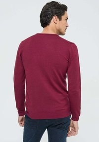 Pull marron à manches longues, avec un col en côtes et un tissu texturé. Porté avec un jean foncé, montrant une silhouette ajustée de dos.
