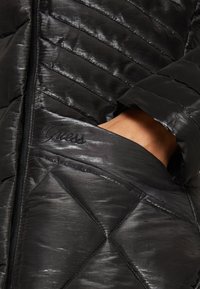 Giacca nera trapuntata con finitura lucida, dotata di chiusura con zip e logo "Guess" ricamato sulla tasca, con cuciture diagonali.