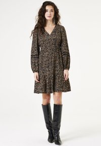 Vestido negro de manga larga con un patrón floral dorado y beige; escote en V, cintura ajustada y falda acampanada; combinado con botas negras hasta la rodilla.