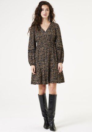 Zwart lange broekjurk met gouden en beige bloemenpatroon; V-hals, getailleerde taille, uitlopende rok; gecombineerd met knielange zwarte laarzen.