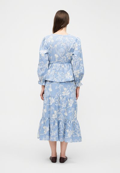 Kvinde med langt brunt hår iført en lyseblå, langærmet kjole med hvidt blomstermønster og konkyliebroderi, stående med ryggen til.