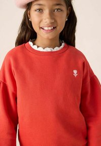 Sweatshirt rouge avec des manches bouffantes, présentant un col blanc en forme de feston et une petite broderie de fleur rose sur la poitrine. Tissu doux.