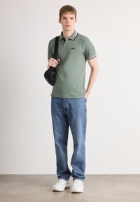 Unge mand iført grøn polo shirt, blå jeans, hvide sneakers og bærende en sort crossbody taske over sin skulder.