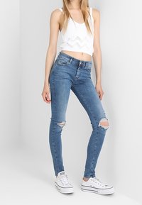 Jeans ajustados azules con roturas desgastadas en las rodillas, de cintura alta y con una textura suave, combinados con un top corto blanco de canalé y zapatillas blancas.