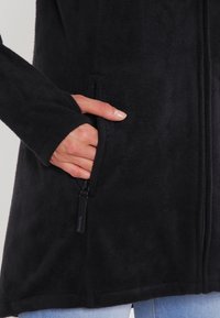 Musta fleece-takki pitkillä hihoilla, jossa on sivupitkä vetoketjulla varustettu tasku ja sileä pinta. Vetoketjassa on musta metallinen viimeistely.