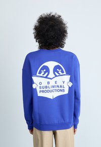 Sweatshirt blu reale in cotone, con una grande grafica bianca che rappresenta due mani che tengono un libro aperto con il testo "OBEY SUBLIMINAL PRODUCTIONS".
