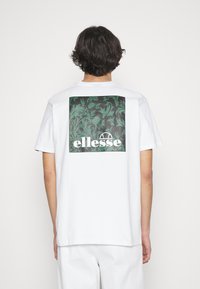 Valge puuvillane t-särk, mille seljal on roheline lillemuster ja brändinimi "ellesse" valgetes tähtedes allosas.