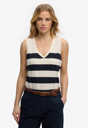 Vrouw draagt een mouwloos wit bovenstuk met brede donkerblauwe horizontale strepen, donkerblauwe broek en een gevlochten bruine riem, staand met een hand in haar zak.