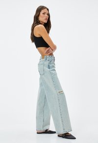 Ljusblå vida jeans med hög midja, slitna detaljer på vänster knä och en läderlapp på bakfickan. Svarta skor.