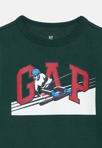 Maglietta GAP verde scuro con scritte rosse e grafica di uno sciatore che indossa un casco e scarponi blu mentre scende in sci.