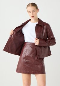 Bordeaux leren jack met een knoopsluiting en grote kraag, gedragen over een witte top. Gecombineerd met een bijpassende high-waisted leren rok.