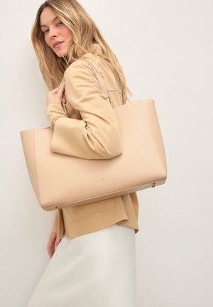 Bež usnjena torba z gladko teksturo, kvadratne oblike in subtilno blagovno znamko; v roki osebe, ki nosi bež zunanji sloj.