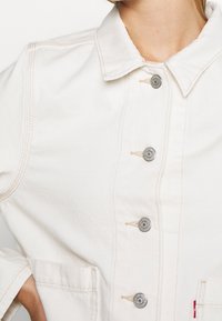 Vit denimjacka med krage, framknäppning med knappar och två stora fickor. Har subtila sömmar och silverfärgade detaljer.