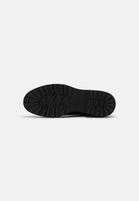 Semelle de botte noire avec un motif de crampons profond et robuste, conçu pour la traction et la durabilité, présenté sur un fond blanc uni.