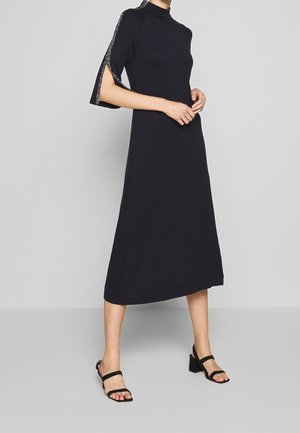 Czarna dzianinowa sukienka z wysokim kołnierzem, rozszerzanymi krótkimi rękawami z srebrnymi akcentami. Sylwetka w kształcie litery A sięgająca do połowy łydki, zestawiona z sandałami na słupkowym obcasie.