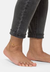 Zlatna anklet s dva privjeska u obliku srca, nosi se na golom stopalu, iznad podvrnutih tamnosivih traperica s vidljivim šavovima.
