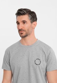 Šedé tričko s krátkým rukávem, vyrobené z měkké látky, s tečkovaným vzorem a kulatým logem s textem. Design s kulatým výstřihem.