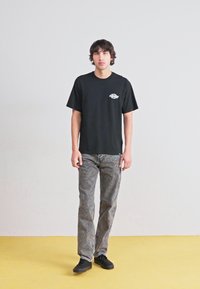 Camiseta de algodón negra con un pequeño logo gráfico en el lado izquierdo del pecho, combinada con unos jeans grises con estampado y zapatos negros.