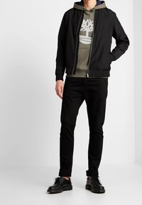Uomo con giacca bomber nera sopra un hoodie oliva con logo bianco ad albero, pantaloni slim neri e scarpe stringate nere su sfondo bianco.