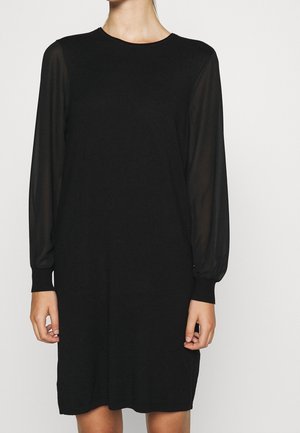 Robe noire longueur genoux avec manches longues transparentes et poignets ajustés, portée par une personne debout sur un fond blanc uni.