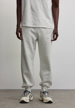 Persona che indossa pantaloni della tuta grigio chiaro, t-shirt grigio chiaro abbinata e sneakers beige, in piedi su un pavimento grigio contro uno sfondo chiaro uniforme.